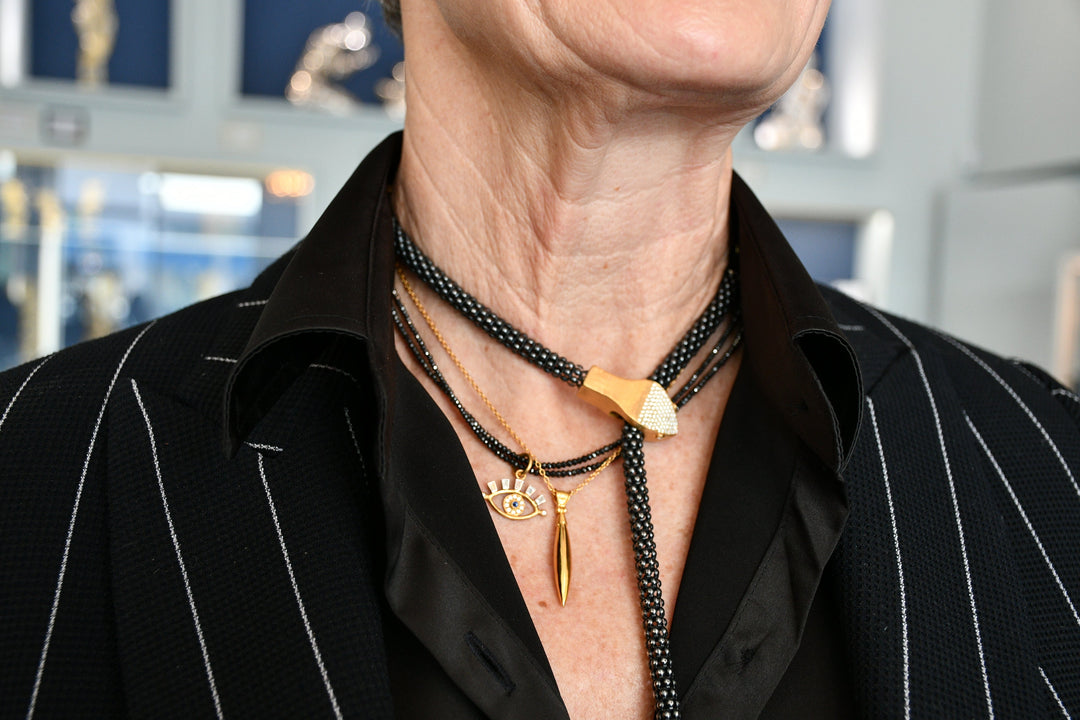 Close-up of a person wearing a black pinstripe jacket and layered black-and-gold necklaces, including an eye-shaped pendant and gold drop charm.