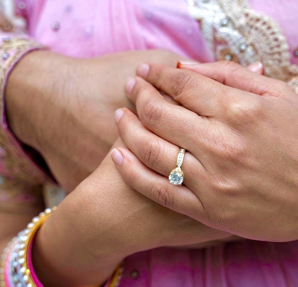 Close-up of a woman’s hands showing a yellow gold engagement ring with a round brilliant diamond, held by her partner while wearing traditional pink and gold attire.