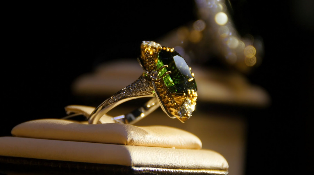 A stunning gold ring featuring a vivid green gemstone surrounded by intricate diamond accents, displayed on a velvet jewelry stand under warm lighting.