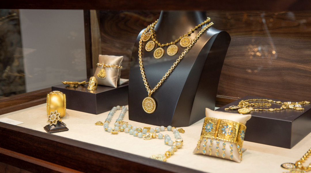 Gold jewelry displayed in a luxury glass showcase with necklaces, bracelets, rings, and ornate pieces arranged on black stands and cream pillows.