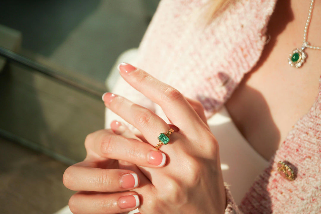 A close-up of a woman’s hands wearing a gold ring set with a rectangular green gemstone, resting gently together. She has a light pink French manicure, and a matching green gemstone necklace is slightly blurred in the background.